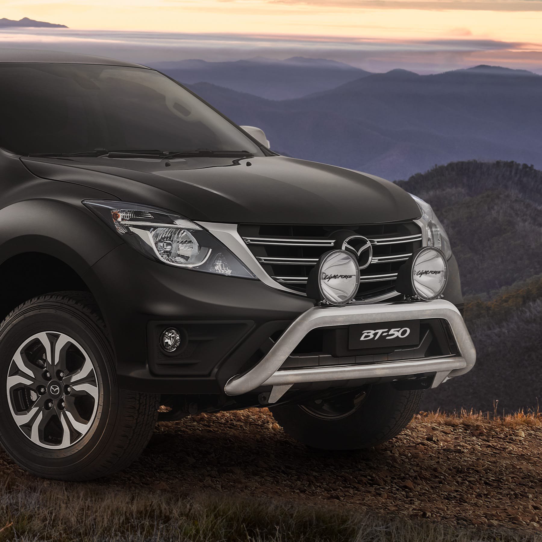 Front-end close-up of a black Mazda BT-50 fitted with a polished nudge bar and driving lights, parked on a ridge at sunset