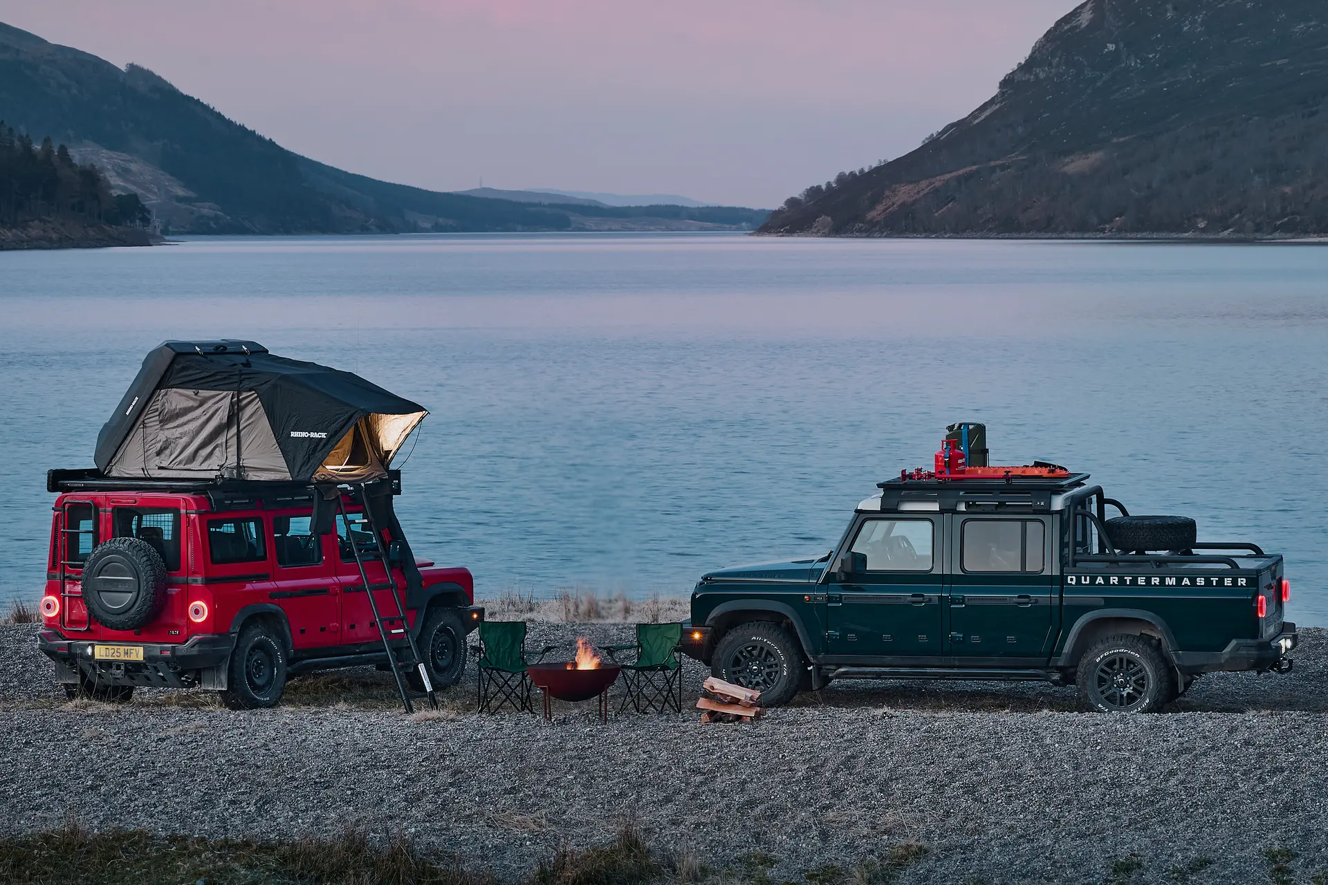 Red INEOS Grenadier Station Wagon with a roof tent beside a green Quartermaster pickup with a roof rack, camped by a loch at dusk