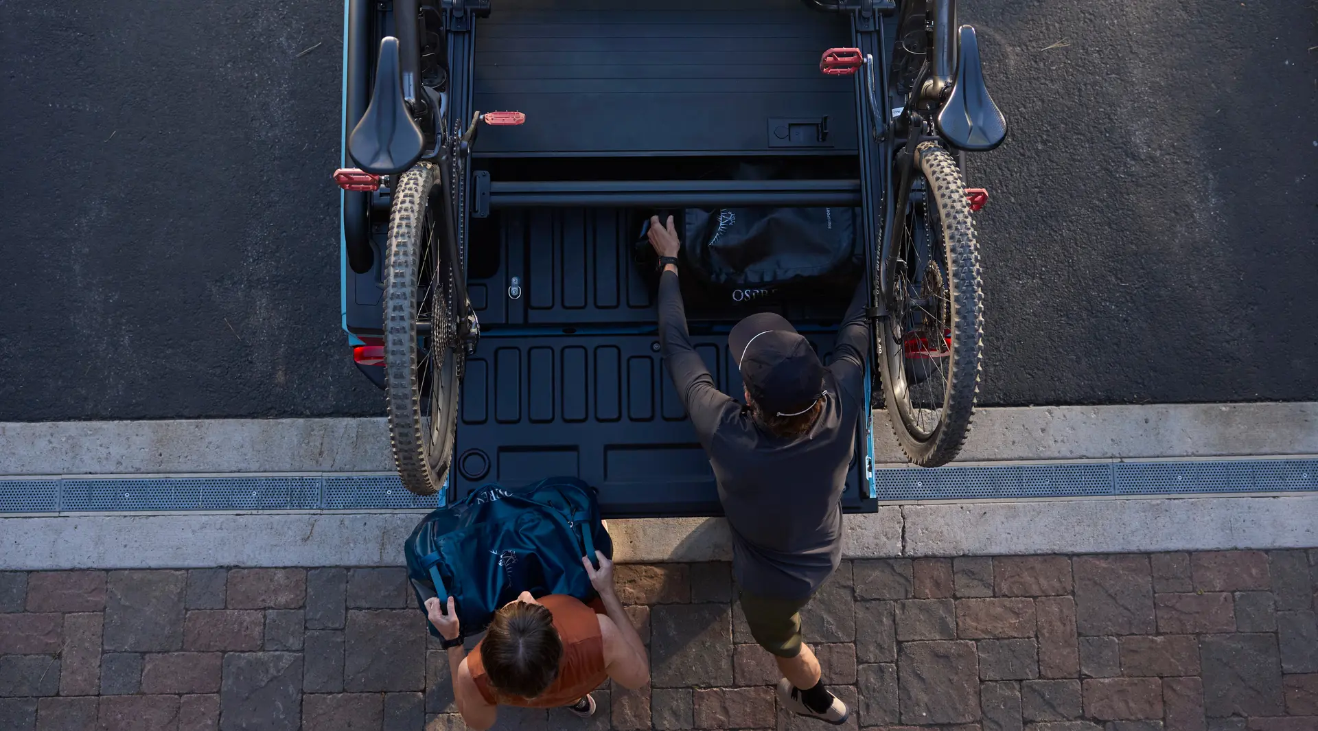 Overhead view of an INEOS Grenadier Quartermaster with the tailgate down, two people loading bikes and kit into the bed