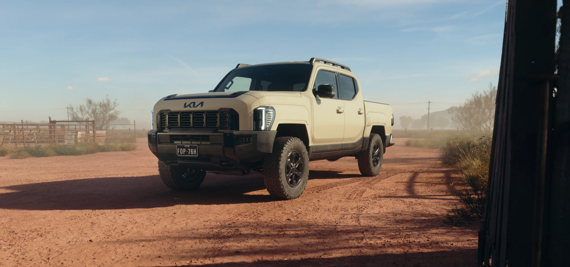 Kia Tasman dual-cab in desert sand parked on red outback dirt beside a stockyard fence, fitted with a front bar and all-terrain tyres
