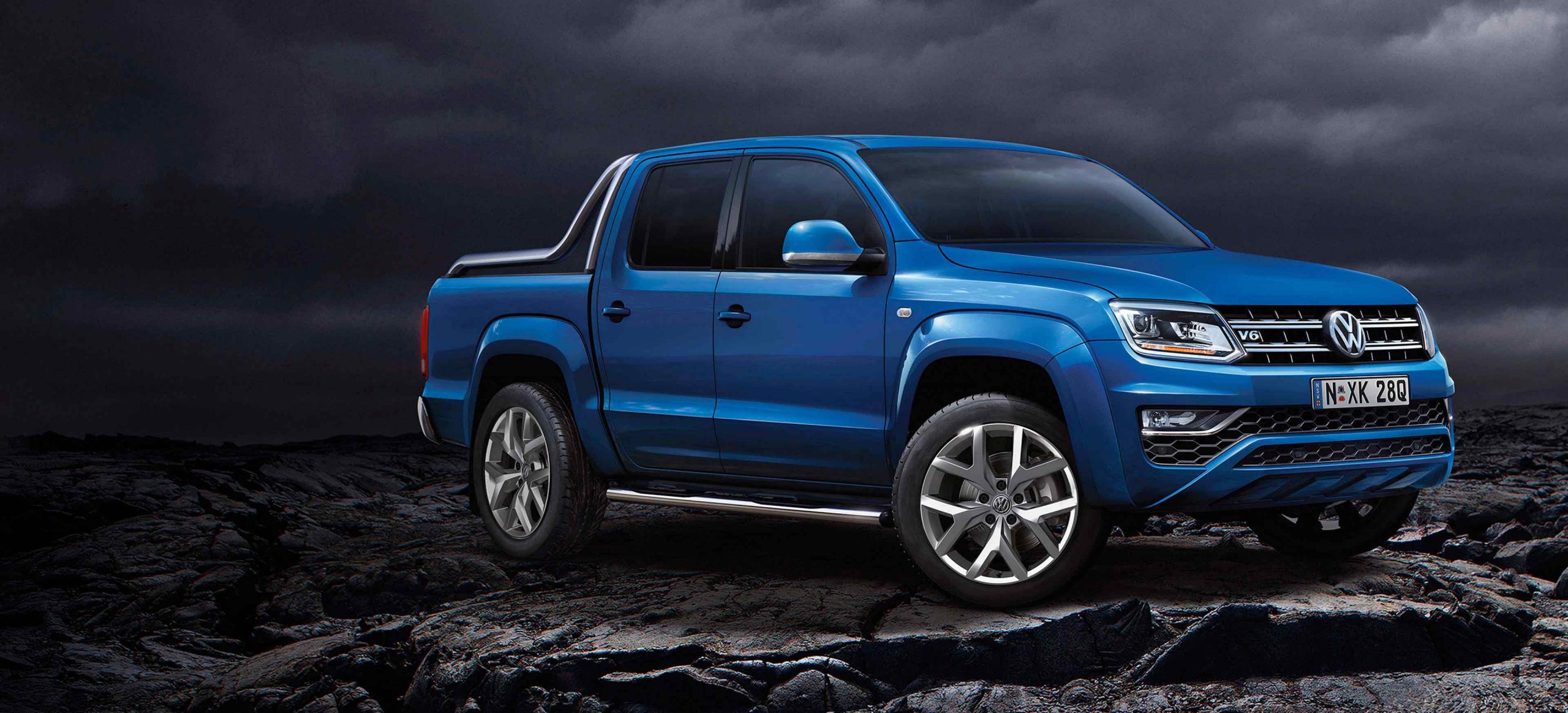 Blue Volkswagen Amarok V6 dual-cab with side steps, parked on dark volcanic rock under a stormy sky